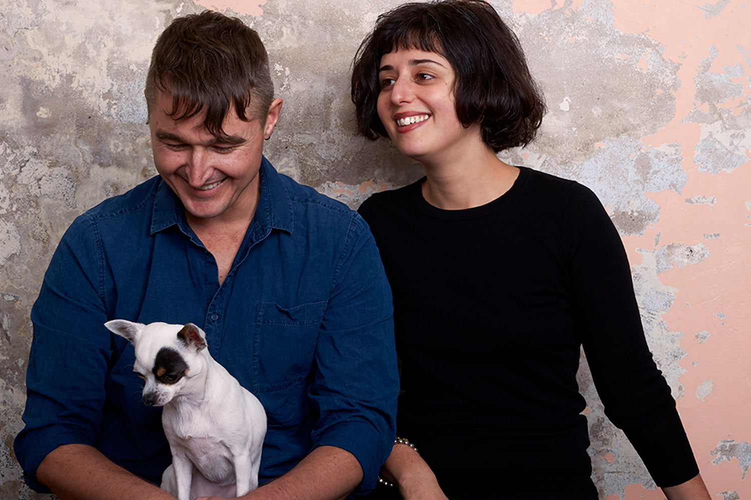 A man and woman both sitting on the ground with a chihuahua, all seated in front of a wall covered in crumbly peach plaster.