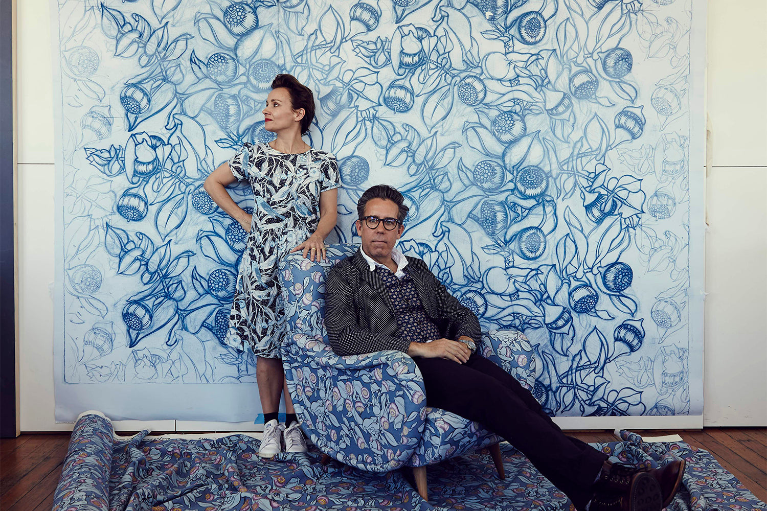 Portrait of a couple in front of a floral backdrop: a man sitting on a patterned chair with his legs spread out, and a woman standing next to the chair.