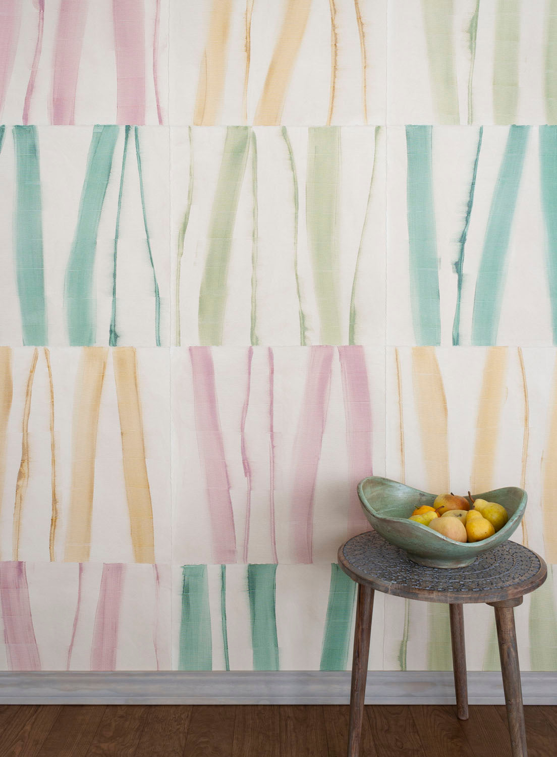A stool stands in front of a wall covered in an irregular striped wallpaper in shades of teal, pink, green and yellow.