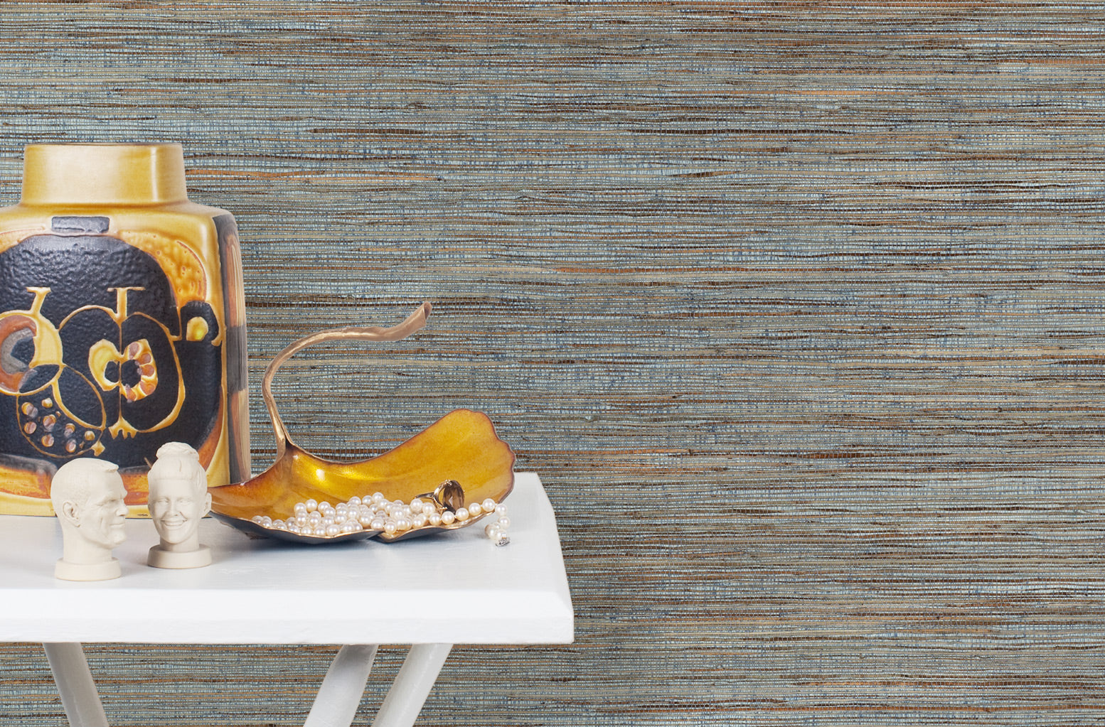 End table with a vase and sculptures in front of a wall papered in jute grasscloth in blue and brown.