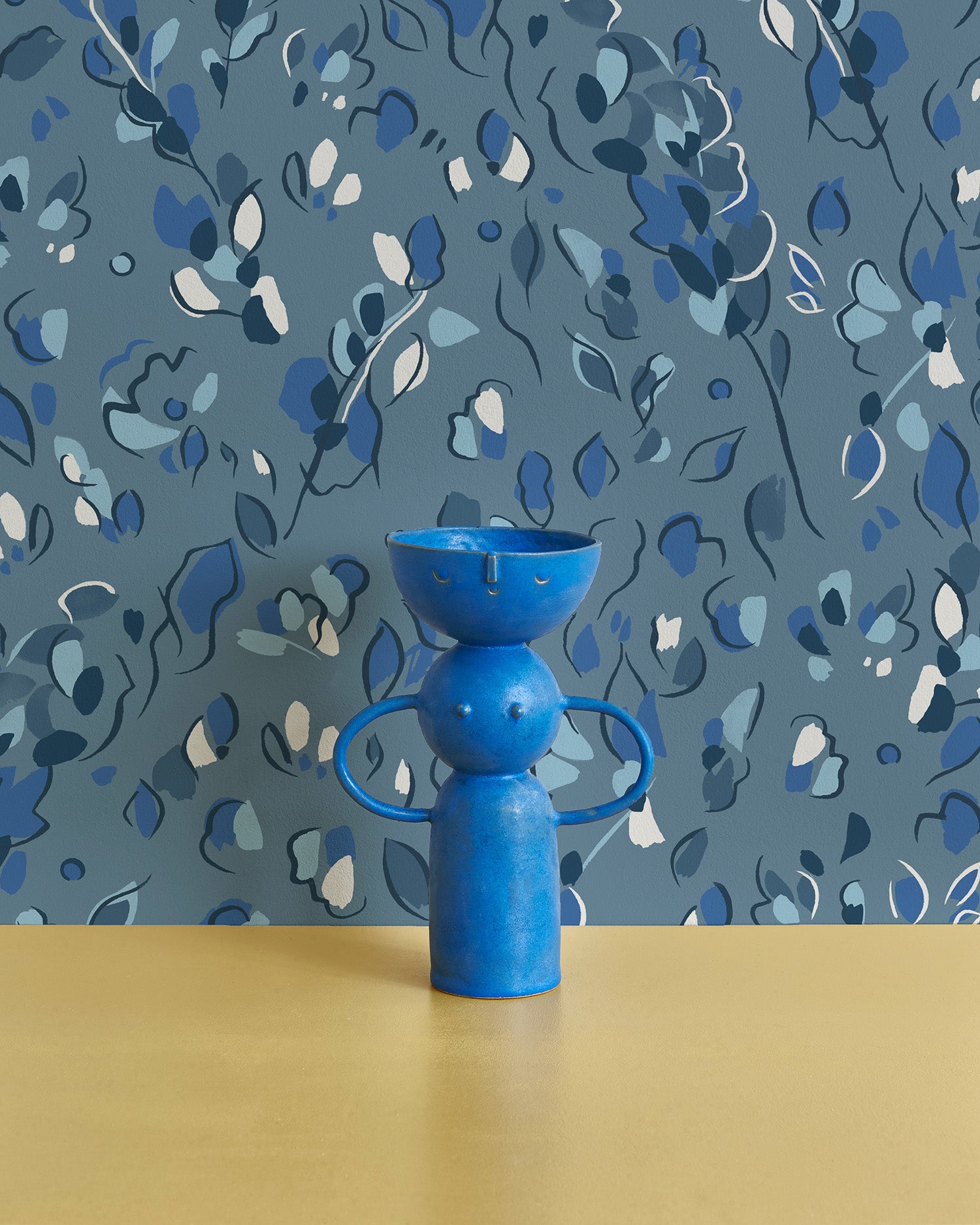 Bright blue vase stands in front of a wall papered in a painterly leaf print in shades of blue.