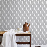 A bench with a towel and clay vases stands in front of a wall papered in an abstract scalloped print in gray and white.