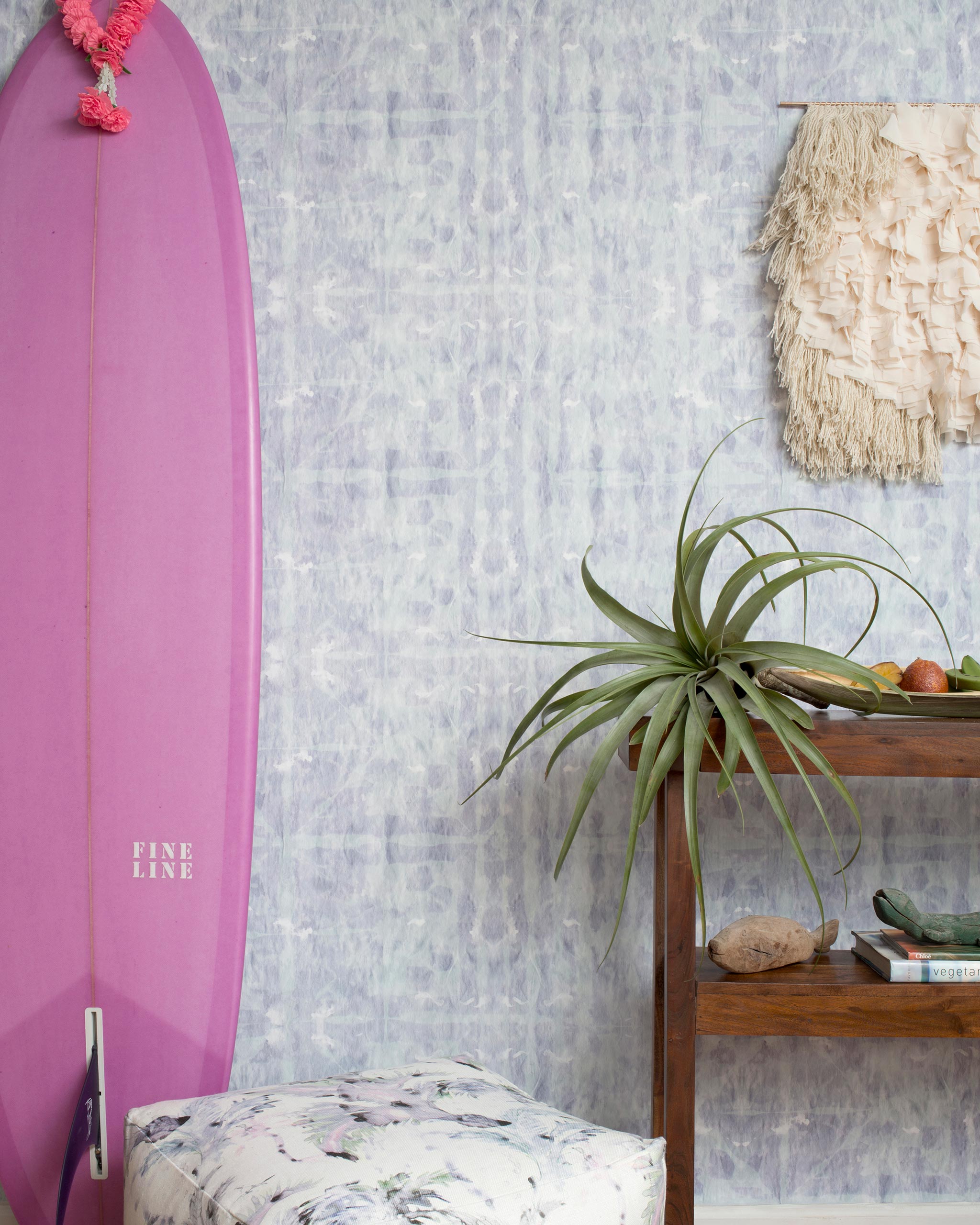 A maximalist room tableau with a purple surfboard and wall papered in an abstract dyed grid print in purple and turquoise.