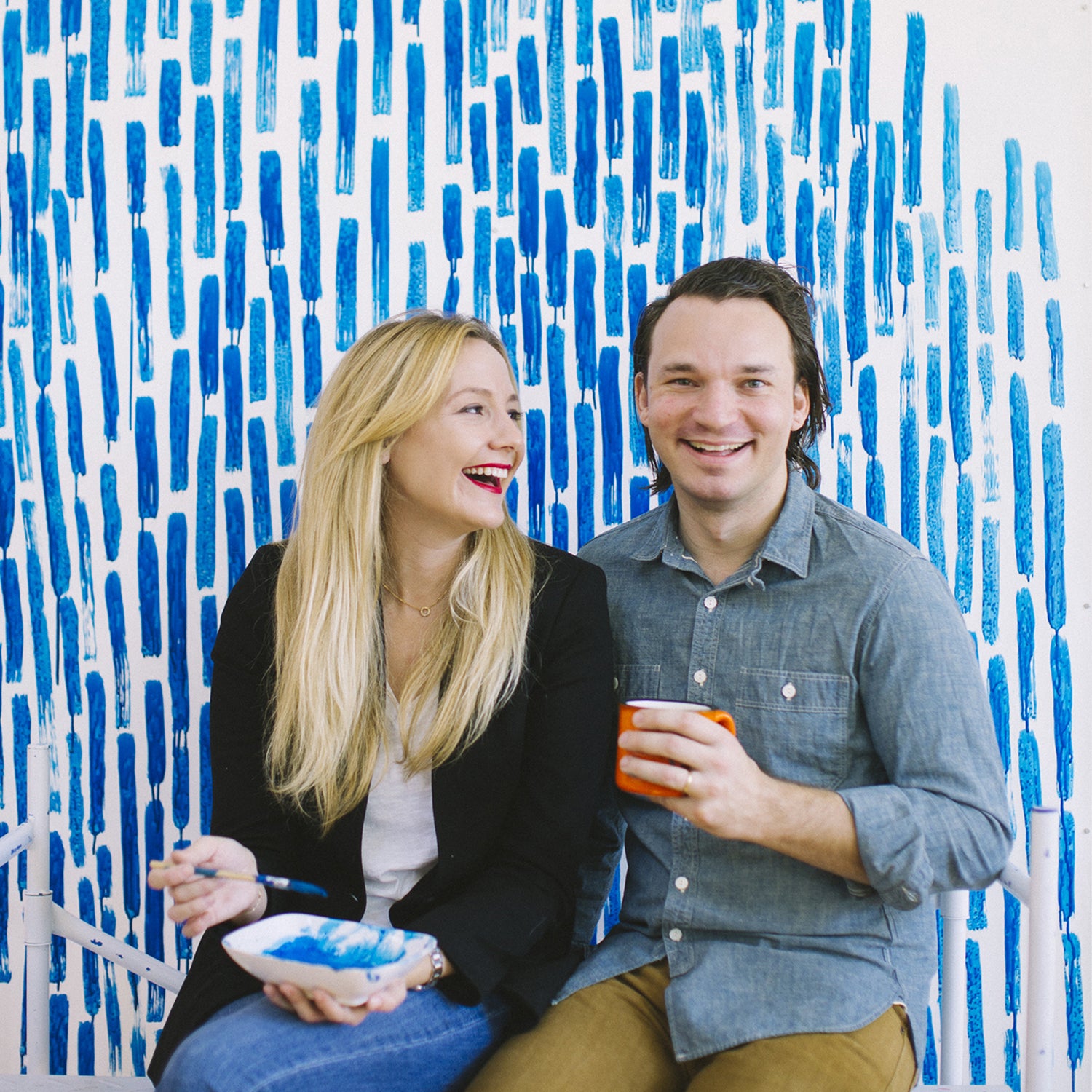 A man and woman sit smiling and holding a paint brush and a coffee mug, in front of a white wall covered in bold blue paint strokes.