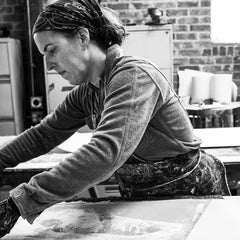 Black and white photo of a woman wearing an apron, long-sleeve button down and bandana working over a screen-printing surface.