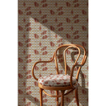 Curved wooden chair with an coral and beige floral cushion in front of a Wallpaper roll with a complex two tone warm red floral motif overlayed a white and tan plaid pattern, hover