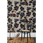 Poppies Noir wallpaper installed on a wall with a chair, lamp and rug, the wallpaper design featuring a black, blue and red floral design with a background of neutral greys and buff tones, hover