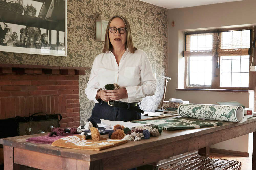 Woman in a white shirt standing beside a wooden table strewn with textile and wallpaper swatches.