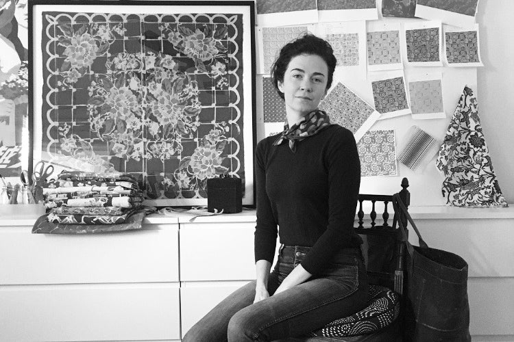 Black and white portrait of a brunette woman sitting on a chair surrounded by a framed textile and many textile swatches.