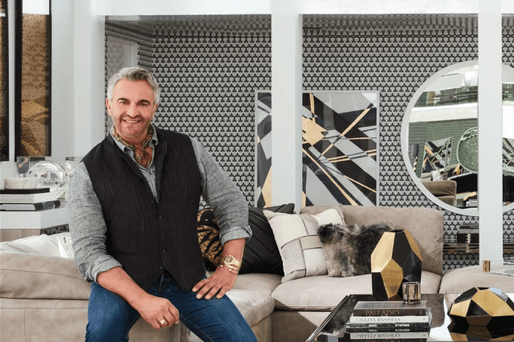 A smiling man seated on a beige couch against a black and white pattered wall. 