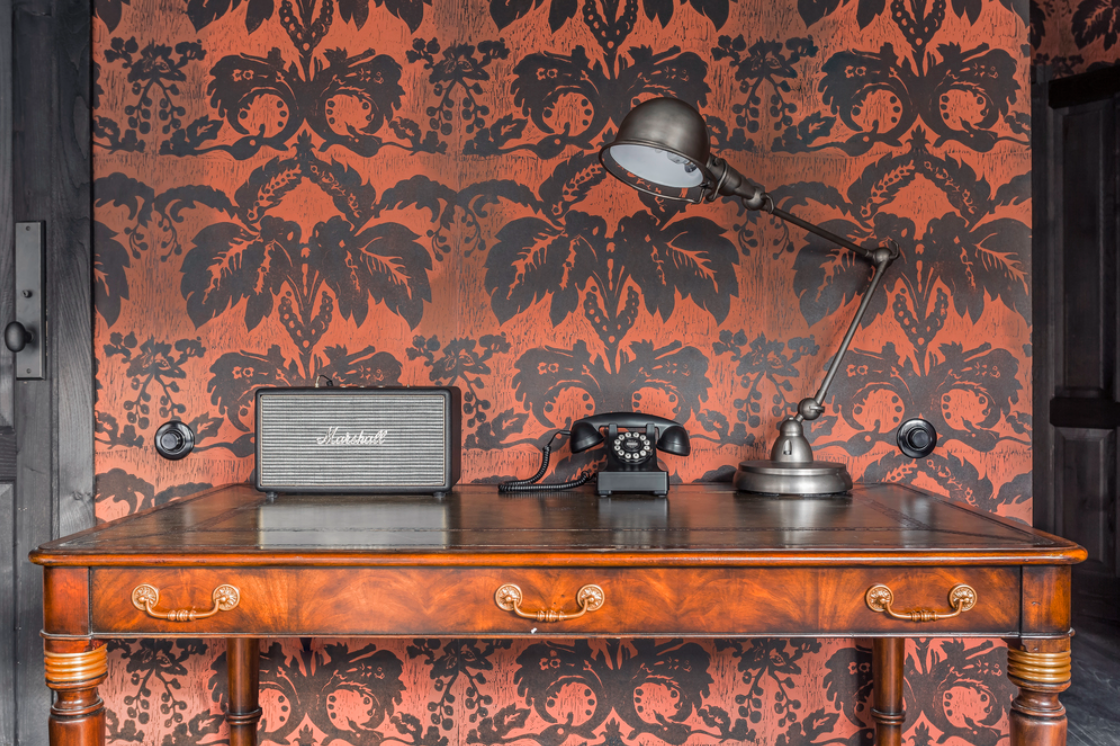 A damask style wallpaper in burnt orange and brown, with a wooden desk in front with vintage office props