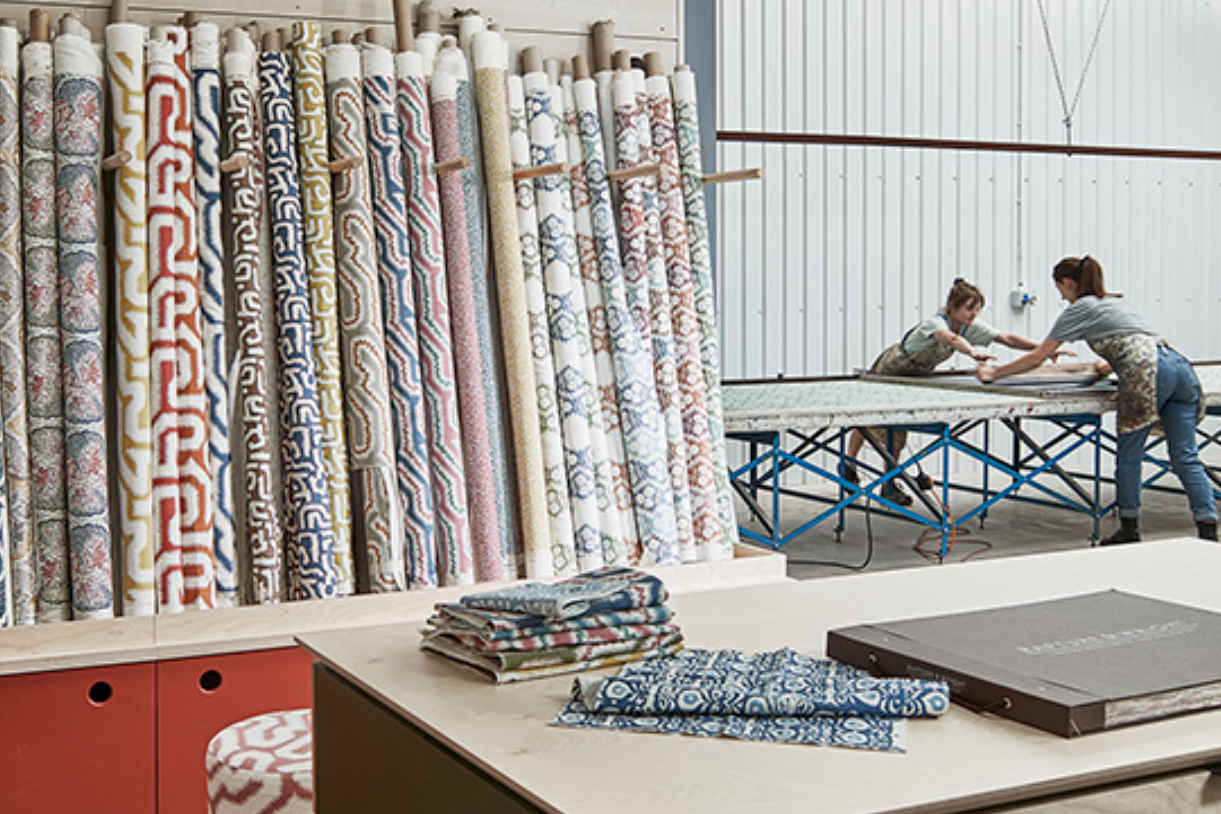 A table with fabric swatches sits in front of a rack of Rtolls of printed fabric, in the background we see two women screen printing at table. 