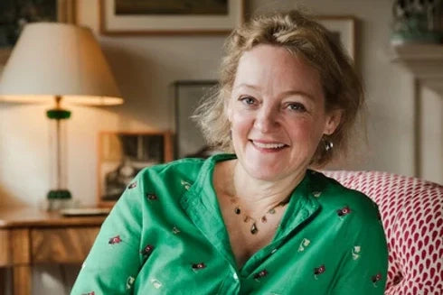 A smiling blond woman in a green shirt, upon a red printed chair in a living room