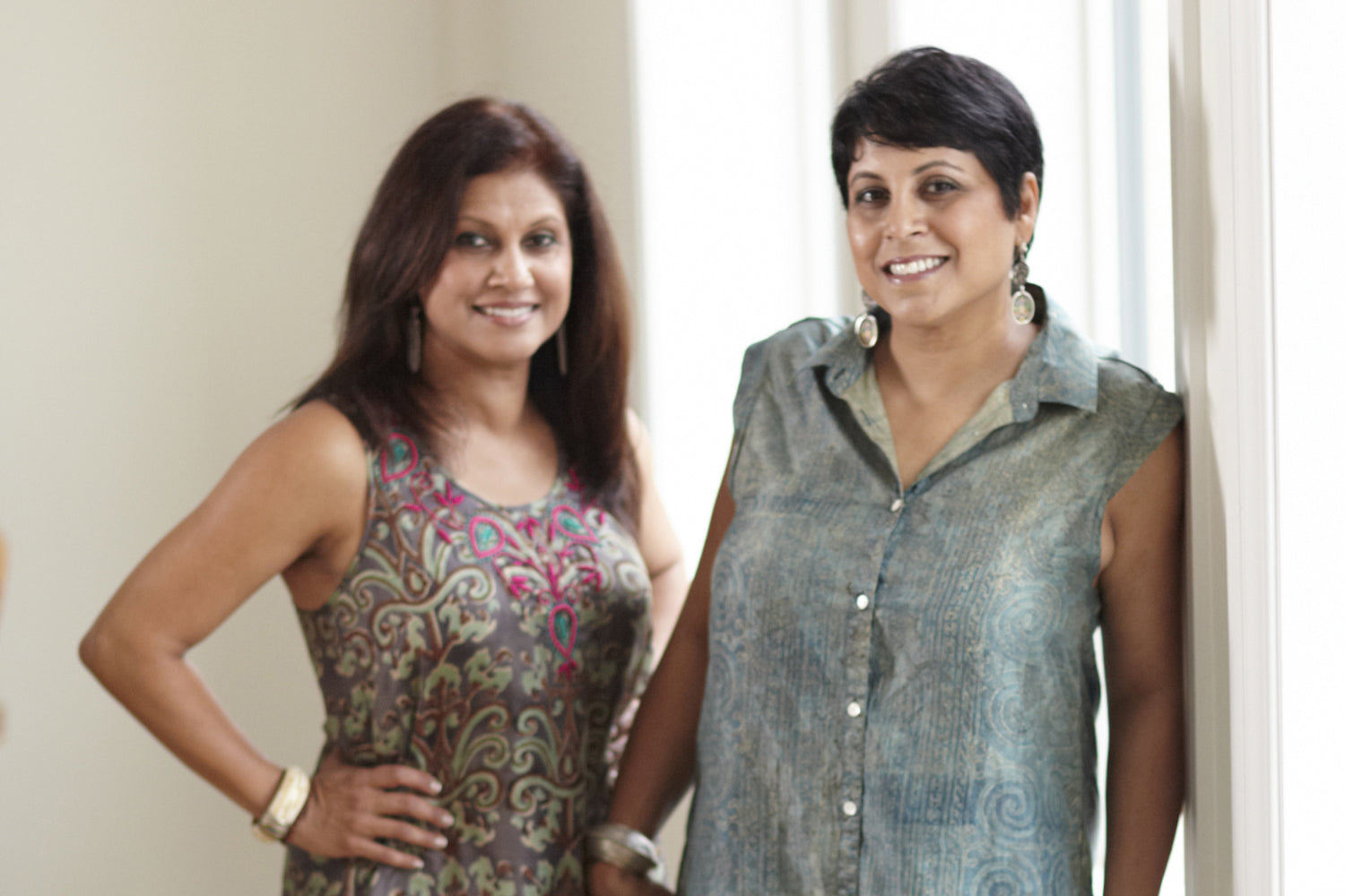 Photo of two brunette women wearing dresses and bangles and standing next to each other in an airy room with white walls. 