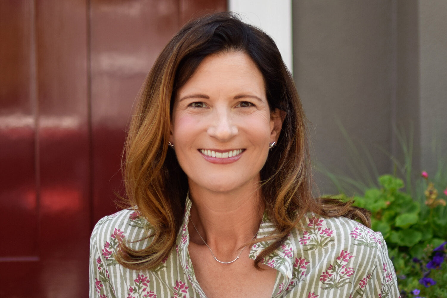 Close-up of a seated woman with straight brown hair, sitting on a front porch in front flowers.