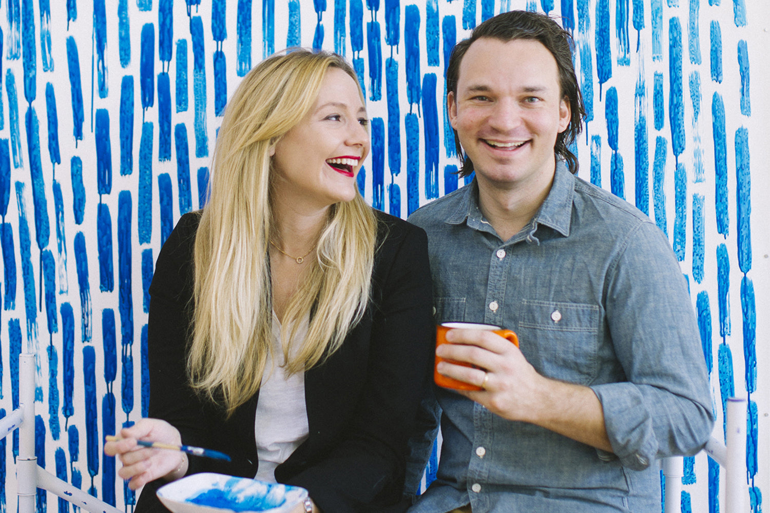 A man and woman sit smiling and holding a paint brush and a coffee mug, in front of a white wall covered in bold blue paint strokes.