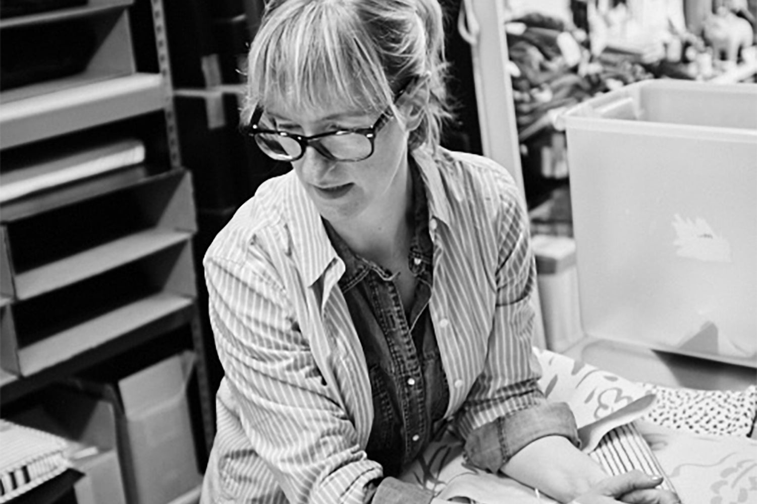 Black and white photo of a woman wearing glasses and a button-down, sitting at a table looking over an array of fabric swatches.