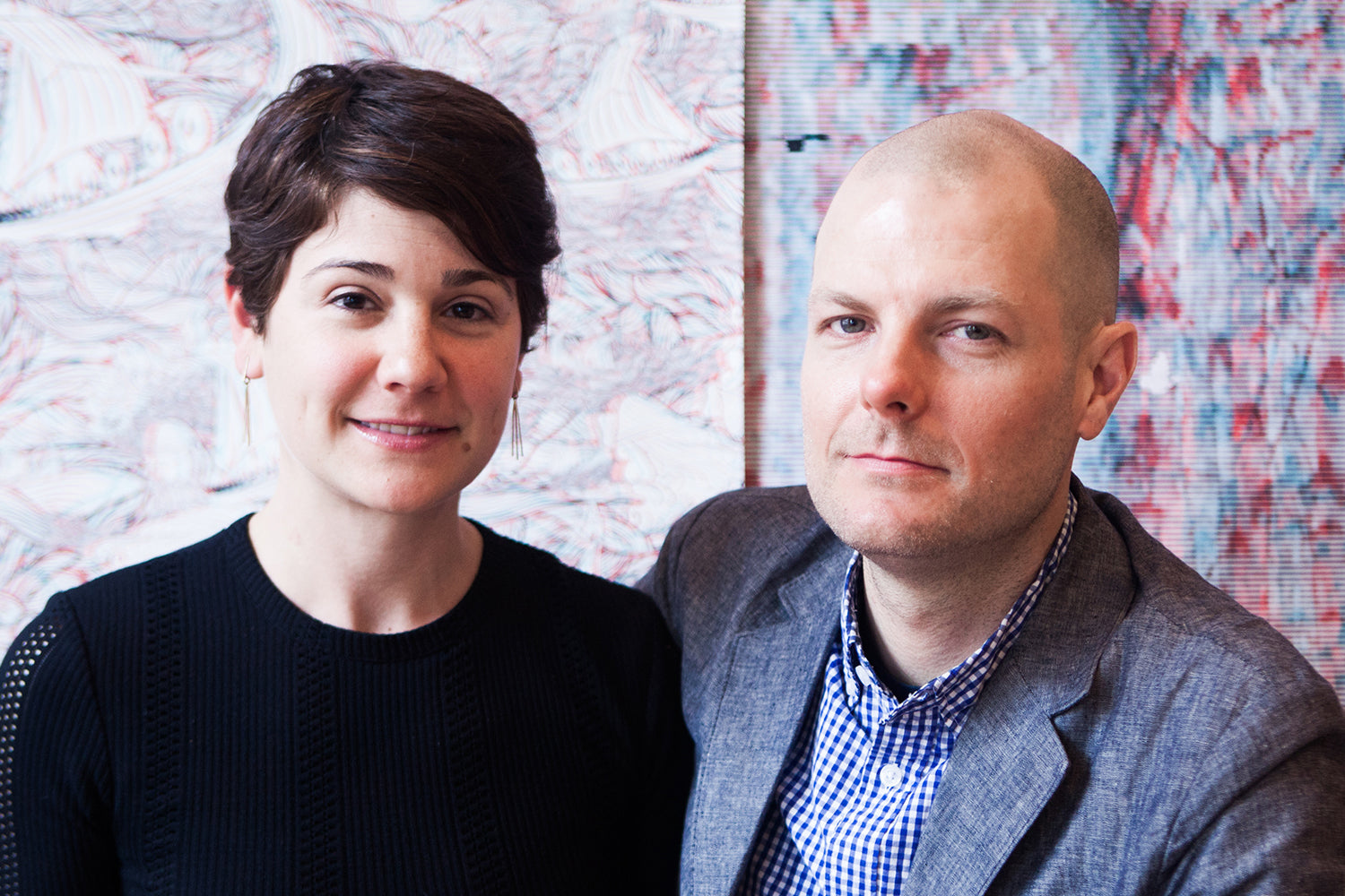 A bald man and a woman with a brunette pixie cut seated in front of a wall papered in two different graphic prints.