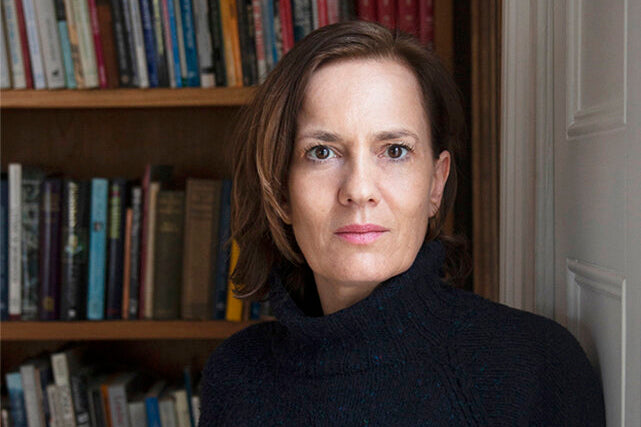 Close-up of a woman with a brunette bob wearing a black turtleneck, seated in front of a wooden bookshelf.