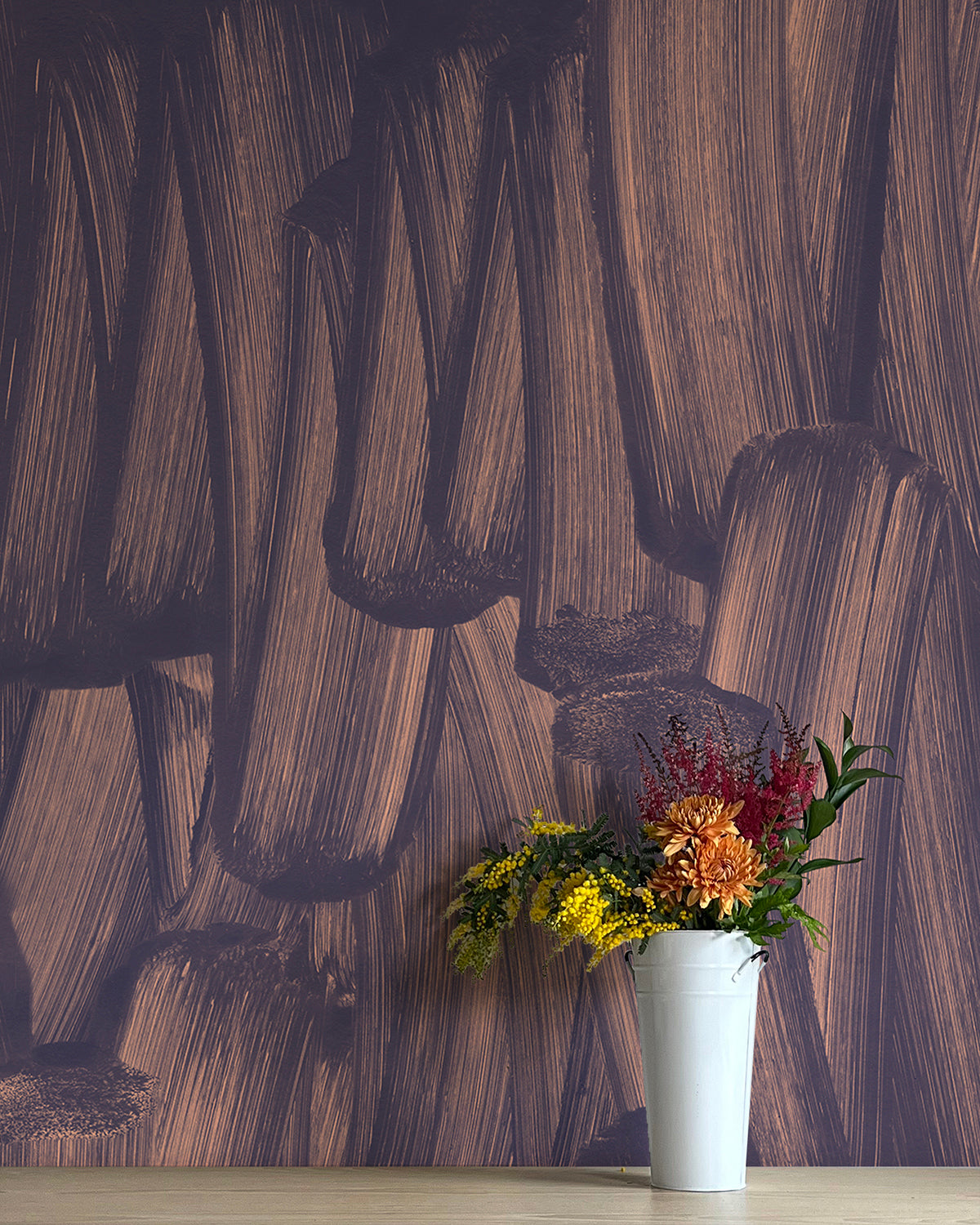 A vase of flowers stands in front of a wall papered in an abstract paint print in dark blue on a purple field.