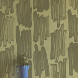 A vase of flowers stands in front of a wall papered in an abstract paint swatch print in dark green on an olive field.