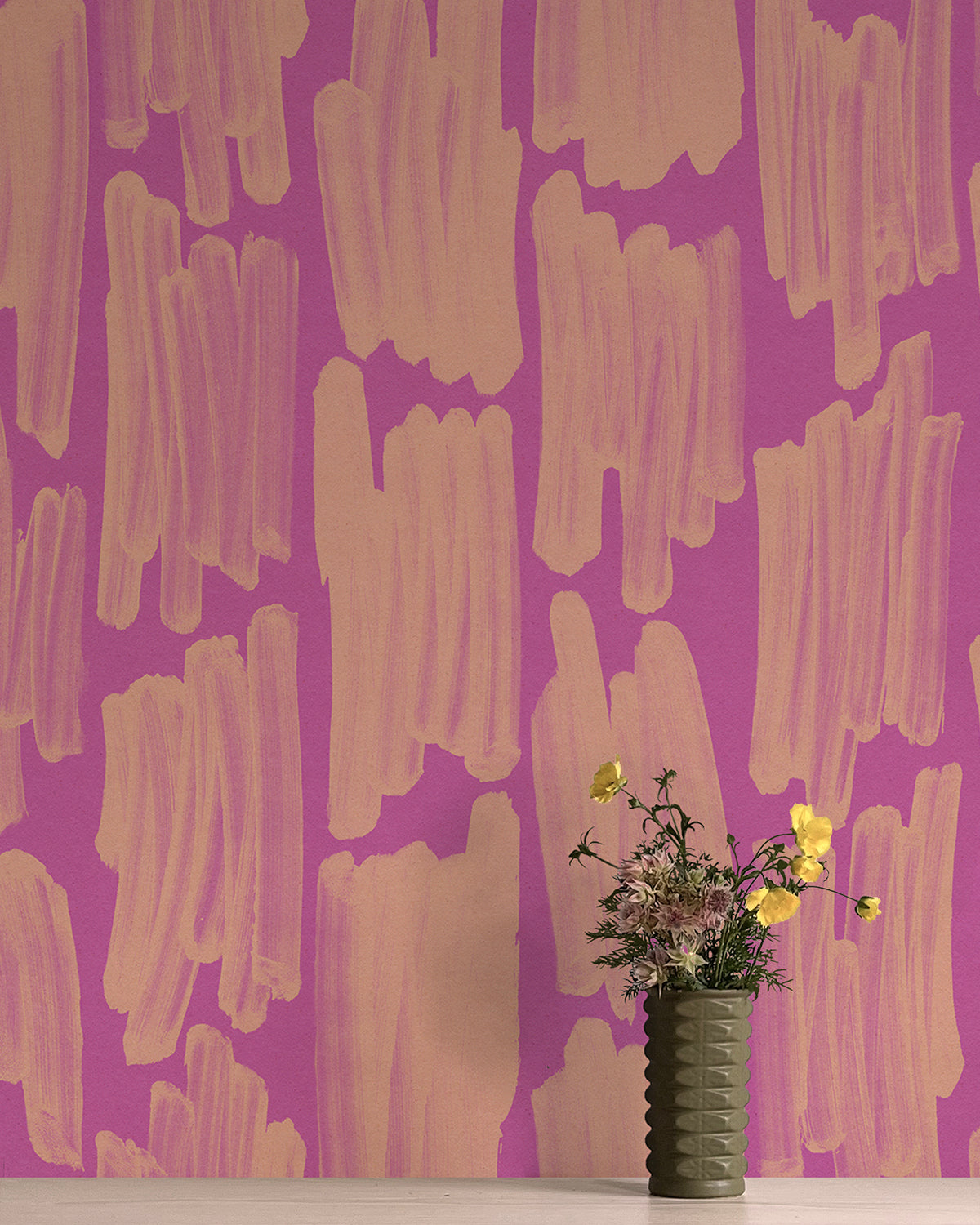 A vase of flowers stands in front of a wall papered in an abstract paint swatch print in orange on a pink field.