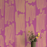 A vase of flowers stands in front of a wall papered in an abstract paint swatch print in orange on a pink field.