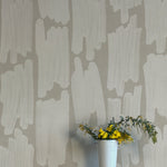 A vase of flowers stands in front of a wall papered in an abstract paint swatch print in light gray on a gray field.