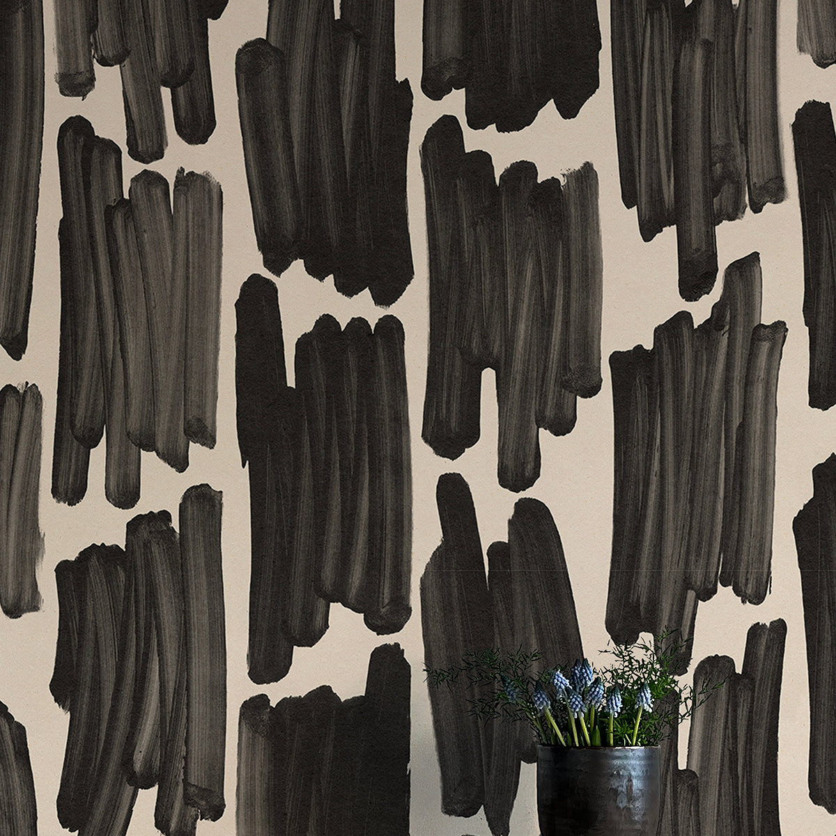 A vase of flowers stands in front of a wall papered in an abstract paint swatch print in black on a cream field.