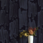 A vase of flowers stands in front of a wall papered in an abstract paint swatch print in blue on a black field.