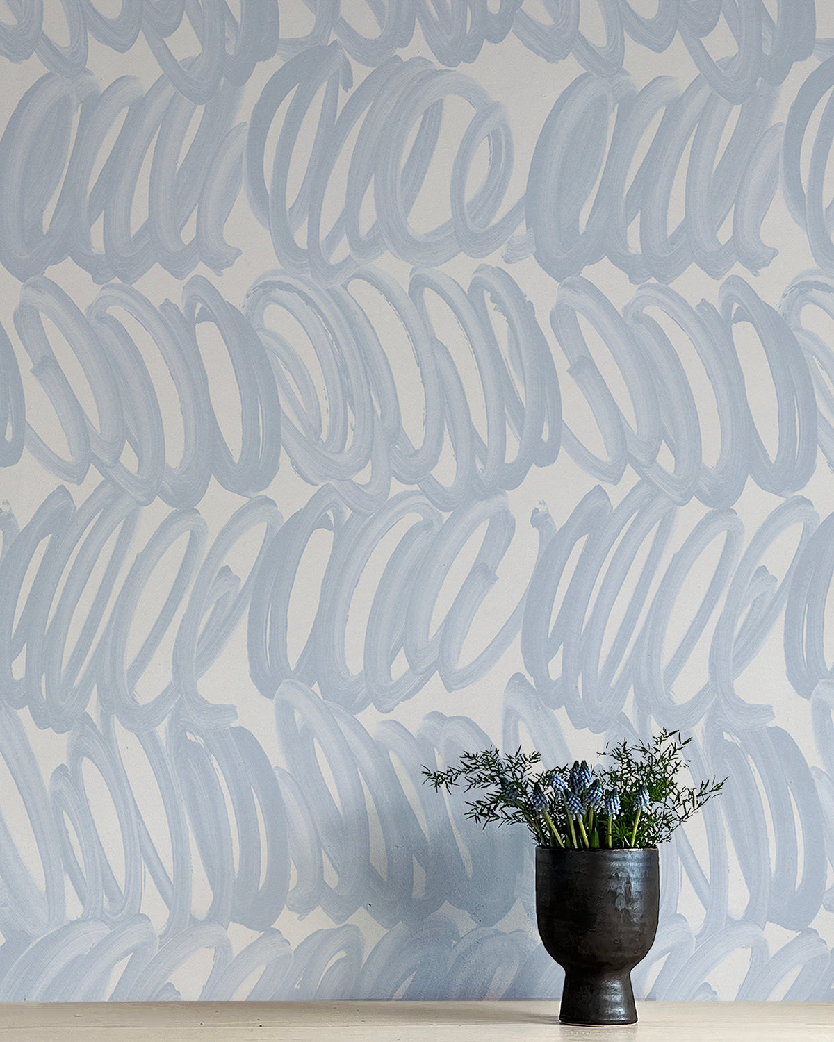 A vase of flowers stands in front of a wall papered in a playful paint print in blue on a light blue field.