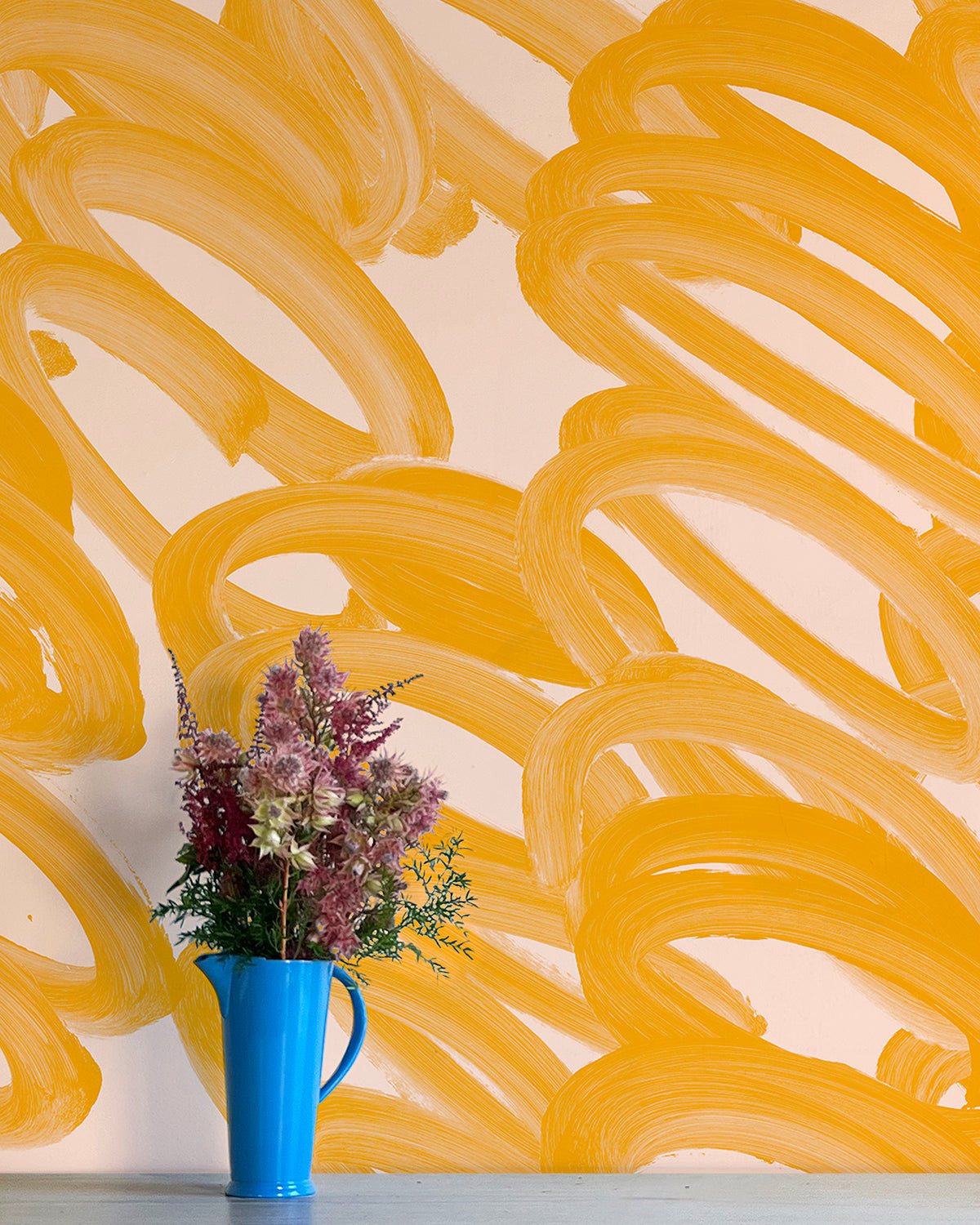 A vase of flowers stands in front of a wall covered in an abstract paintstroke print in orange on a cream field.