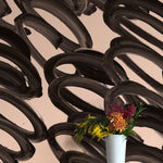 A vase of flowers stands in front of a wall covered in an abstract paintstroke print in black on a cream field.