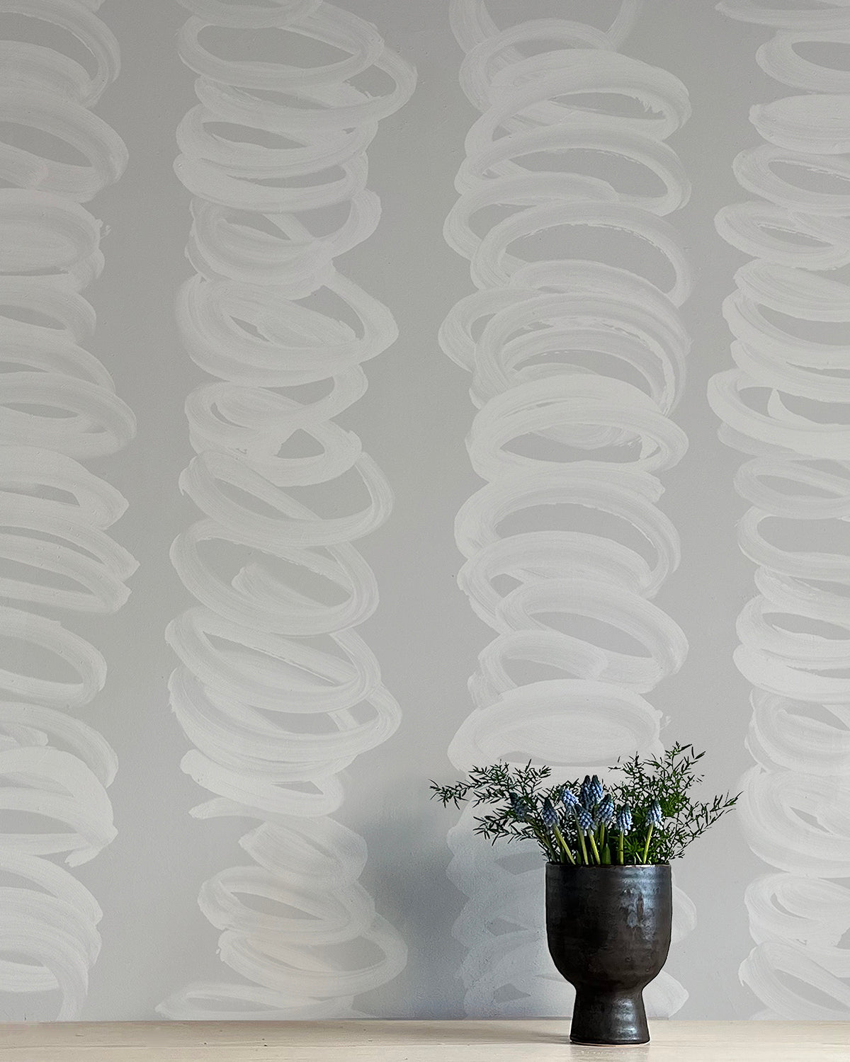 A vase of flowers stands in front of a wall covered in an abstract scribble print in light gray on a gray field.