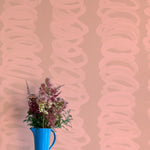 A vase of flowers stands in front of a wall covered in an abstract scribble print in light pink on a tan field.