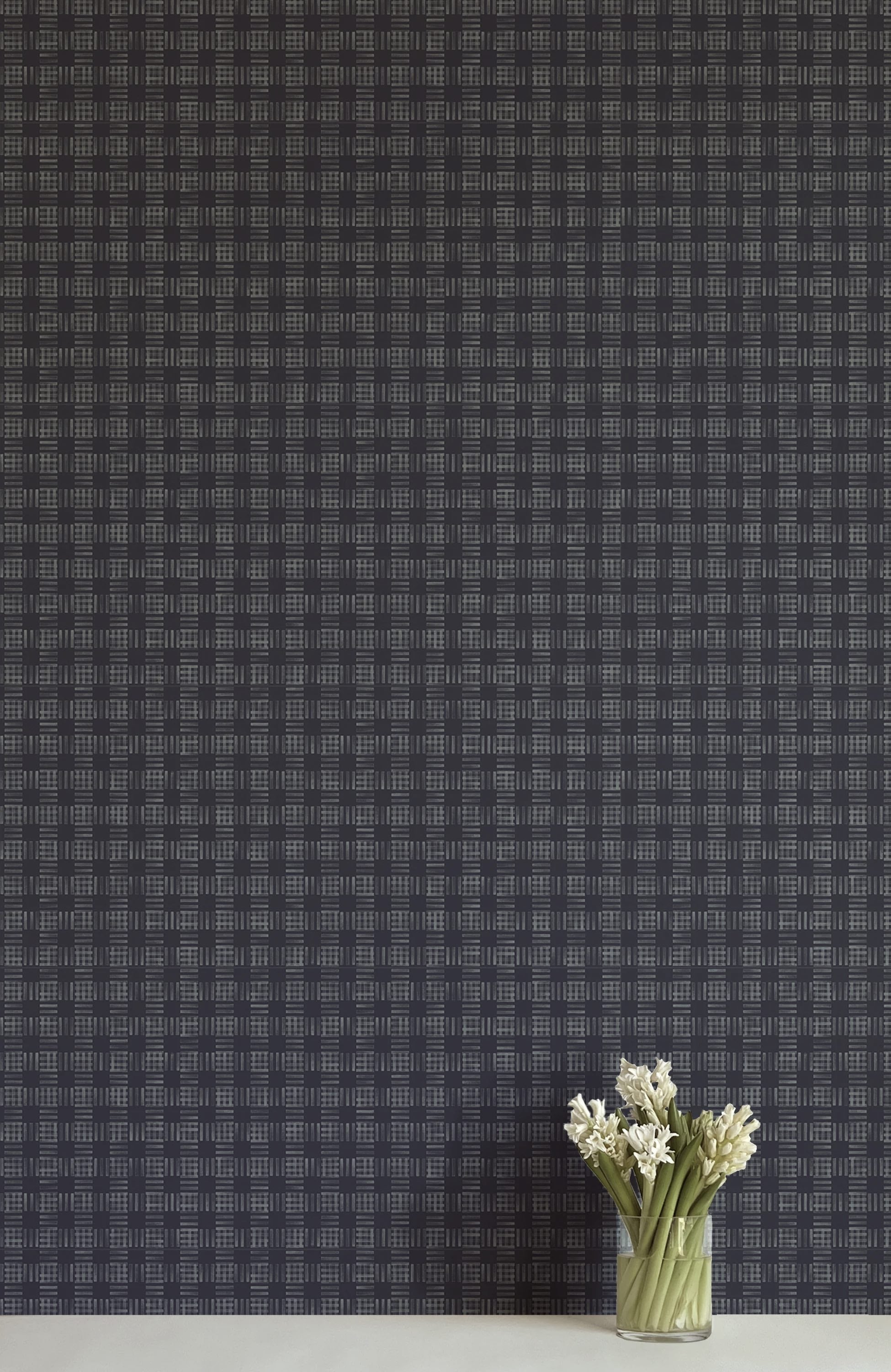 A vase of flowers stands in front of a wall papered in a dense plaid pattern in shades of navy.