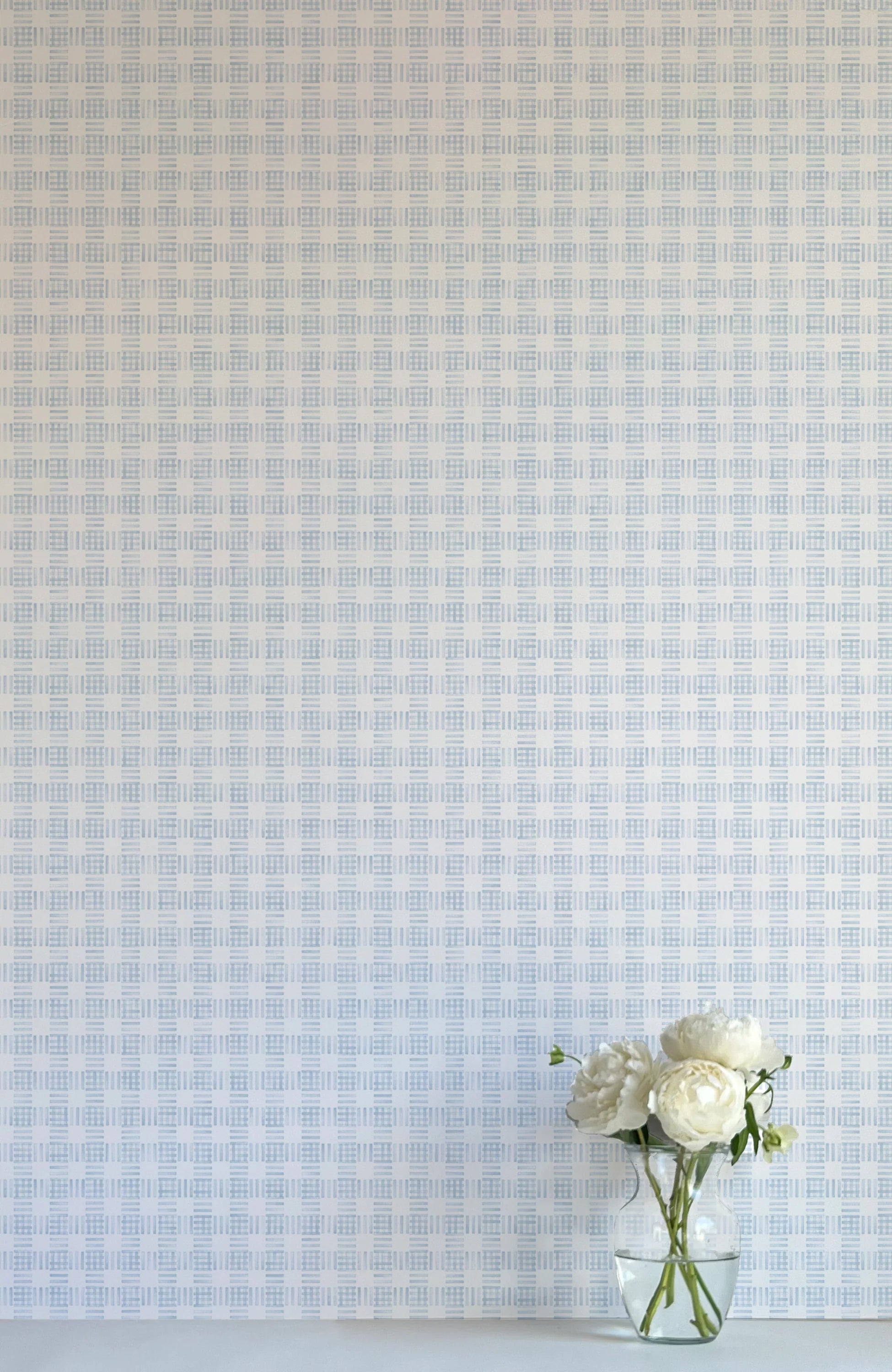 A vase of flowers stands in front of a wall papered in a dense plaid pattern in shades of light blue and cream.