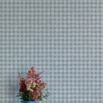 A vase of flowers stands in front of a wall papered in a dense plaid pattern in shades of blue-gray.