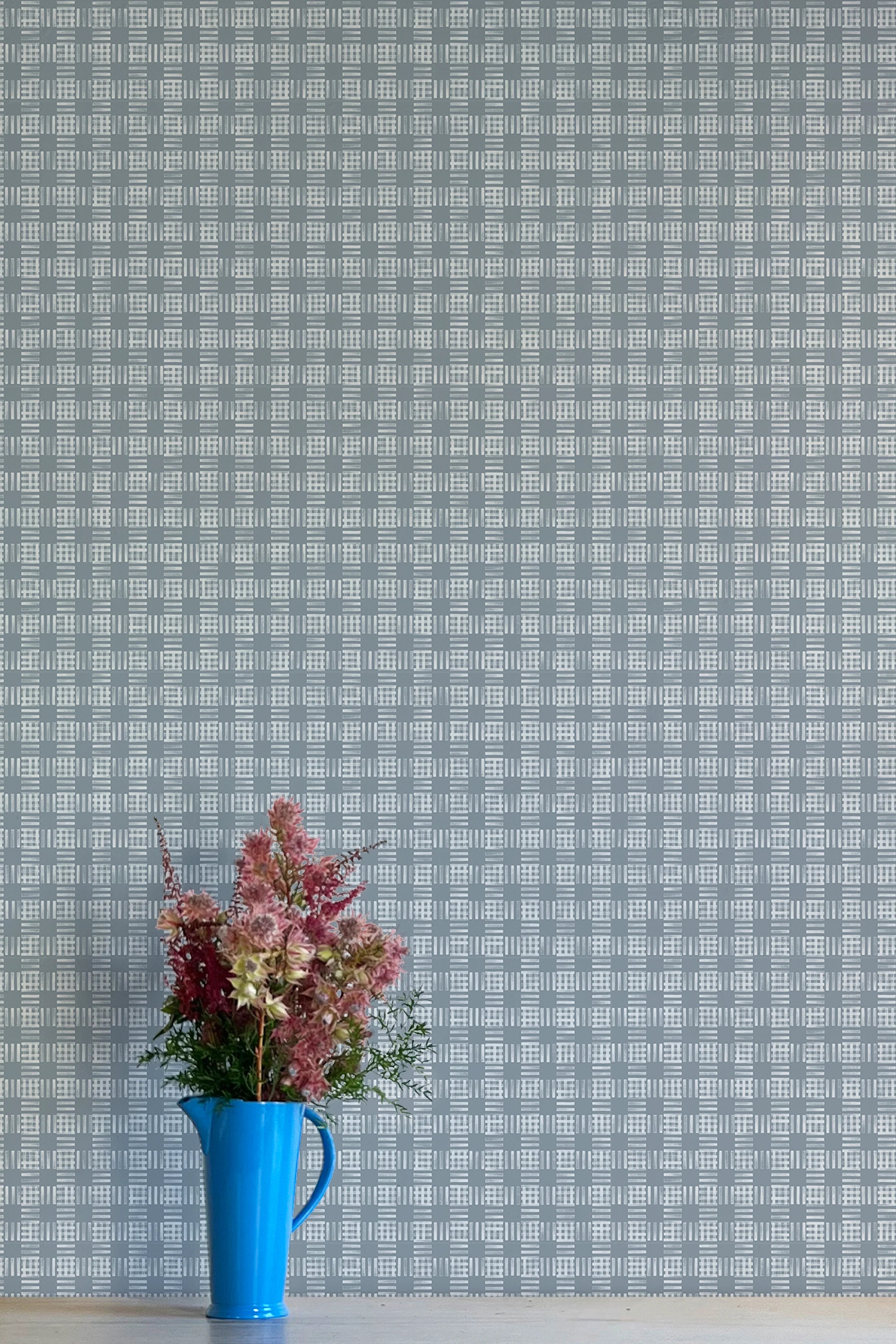 A vase of flowers stands in front of a wall papered in a dense plaid pattern in shades of blue-gray.