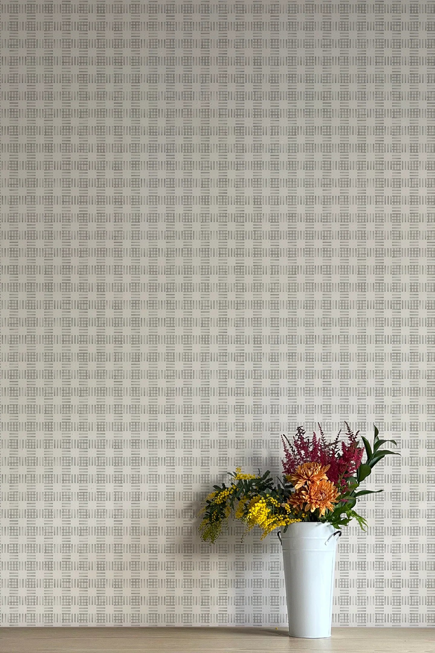 A vase of flowers stands in front of a wall papered in a dense plaid pattern in shades of gray.