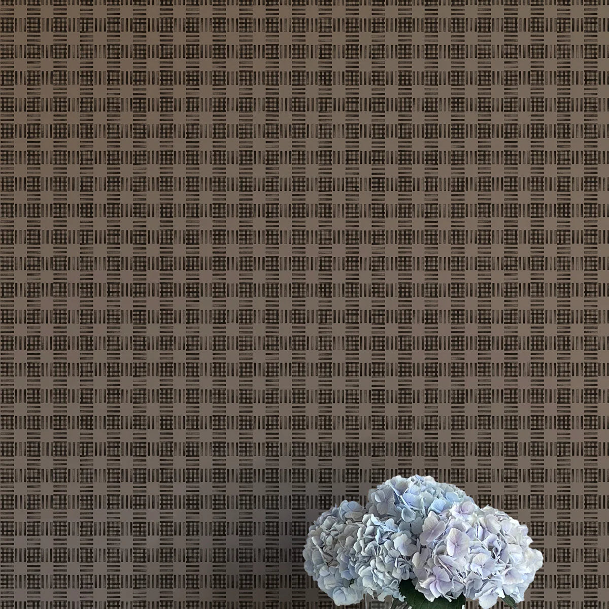 A vase of flowers stands in front of a wall papered in a dense plaid pattern in shades of brown.