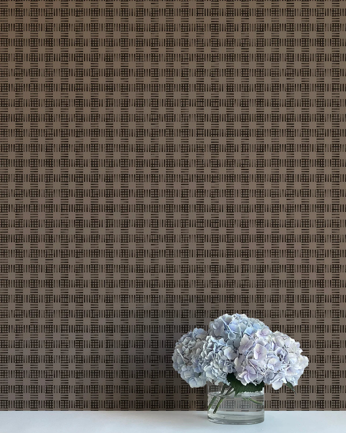 A vase of flowers stands in front of a wall papered in a dense plaid pattern in shades of brown.