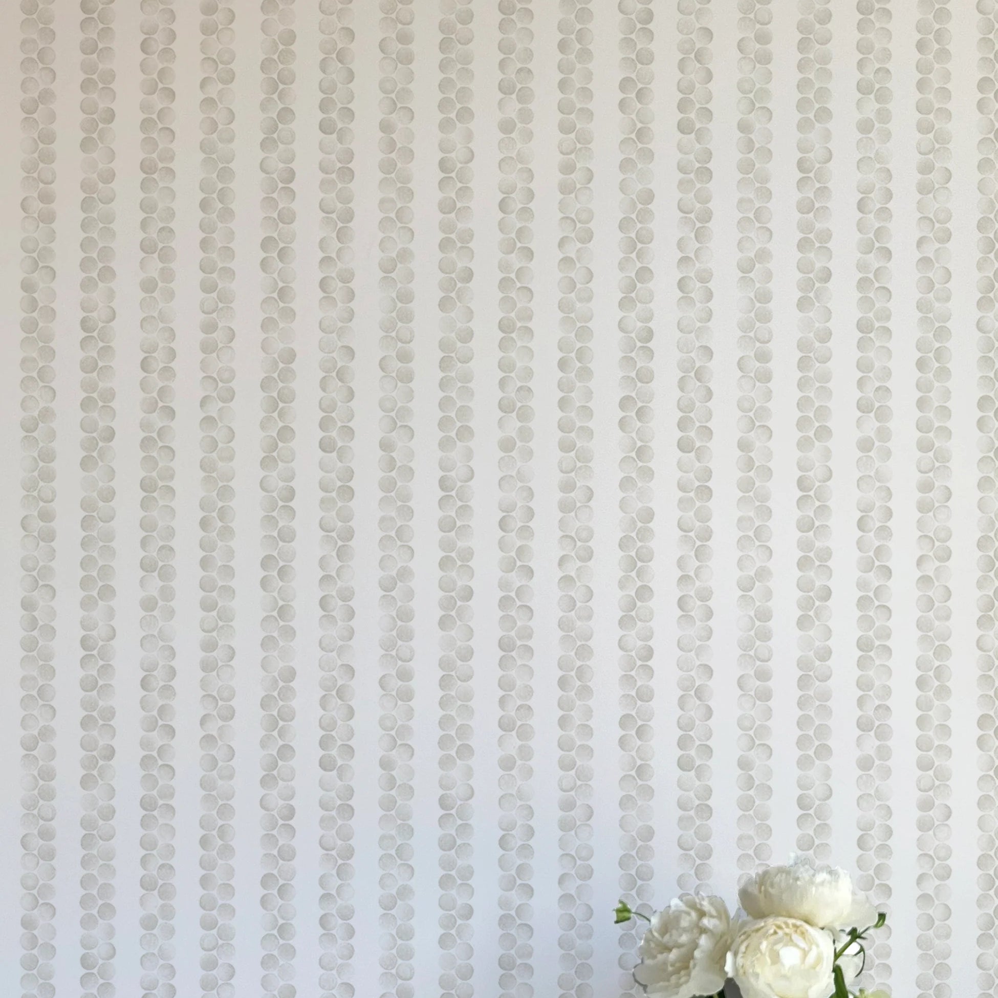 A vase of flowers stands in front of a wall covered in a repeating striped circular print in gray on a light gray field.