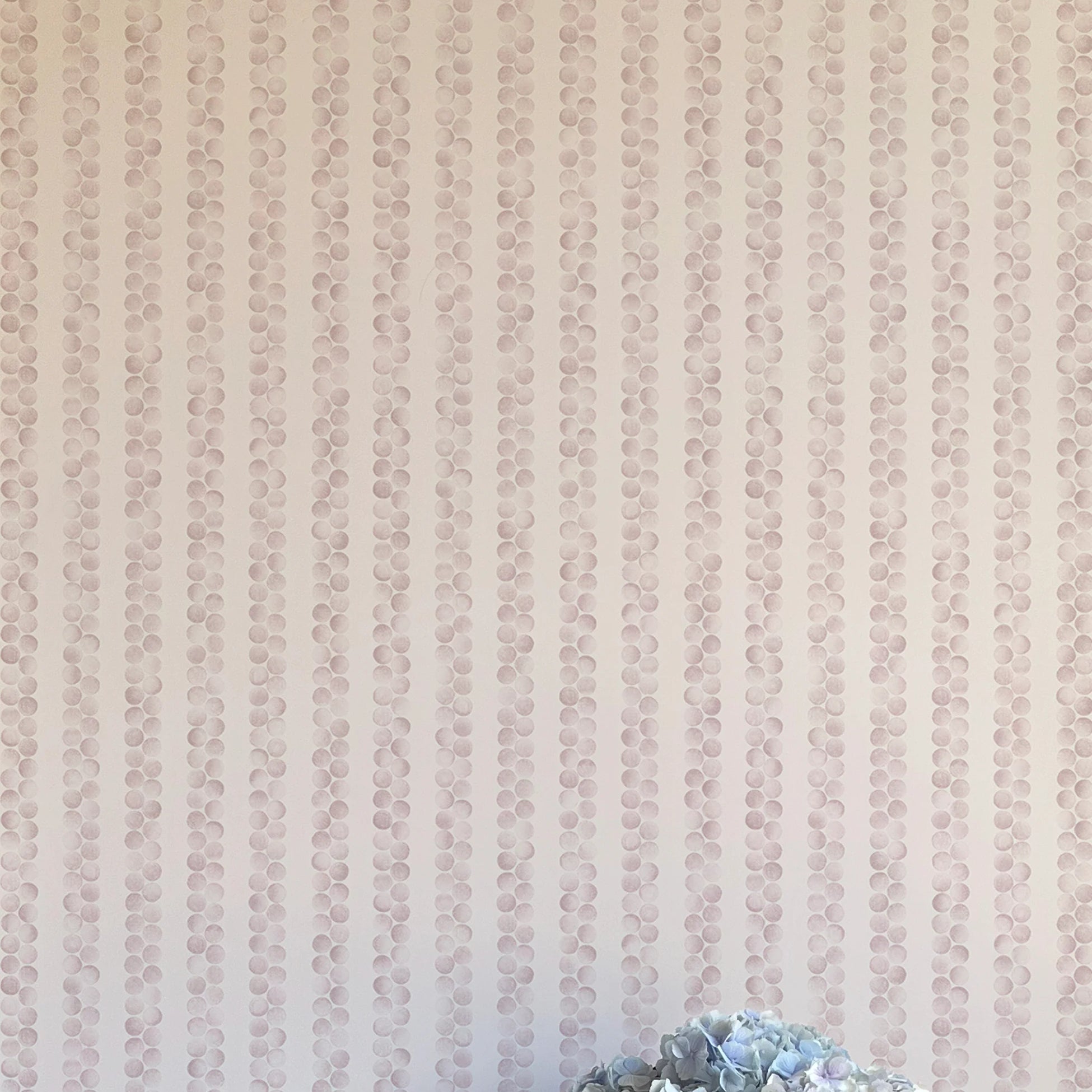 A vase of flowers stands in front of a wall covered in a repeating striped circular print in purple on a cream field.