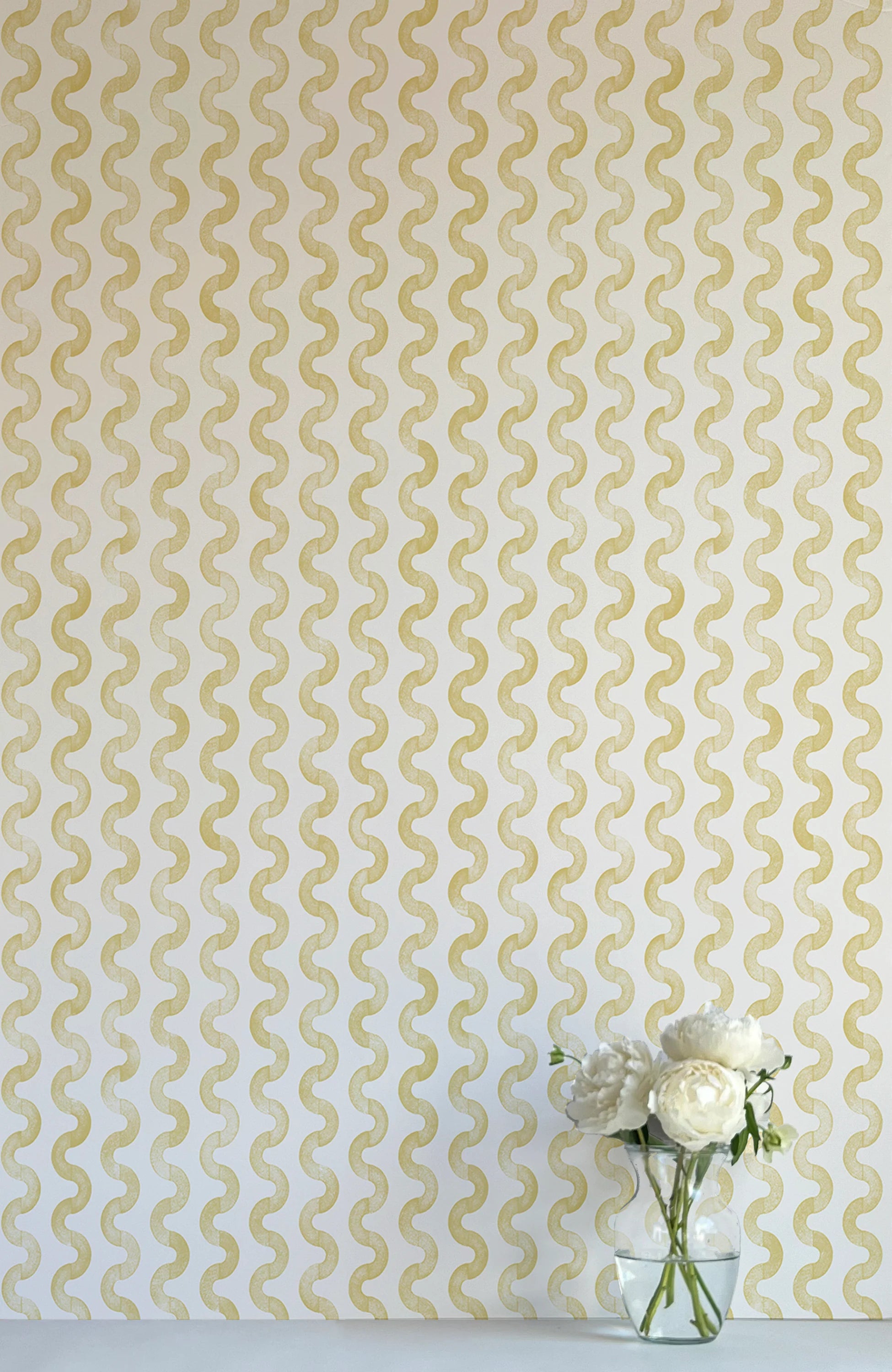 A vase of flowers stands in front of a wall papered in a playful curvy stripe print in yellow on a cream field.