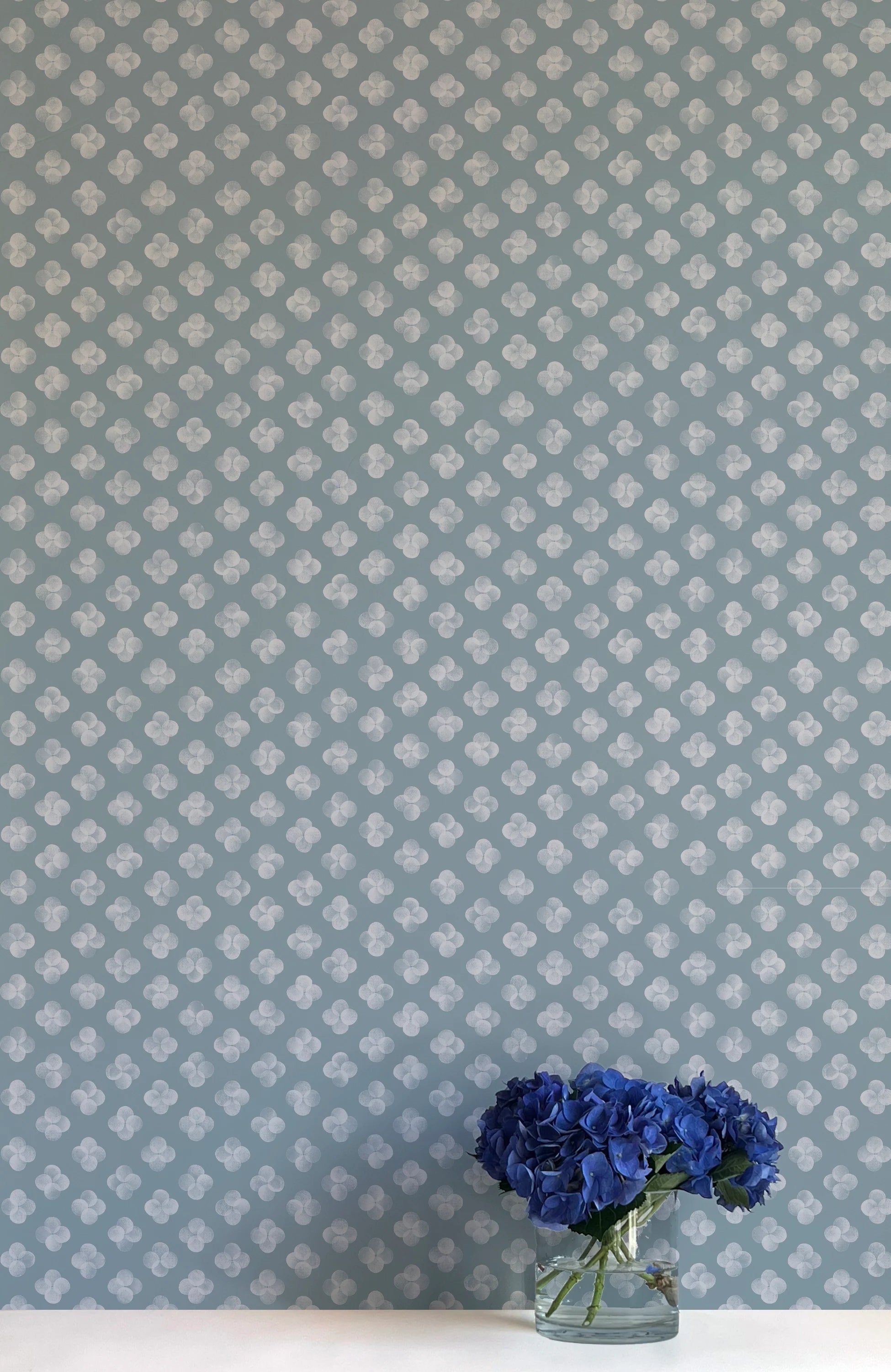 A vase of flowers stands in front of a wall covered in a minimalist quatrefoil pattern in gray on a blue field.