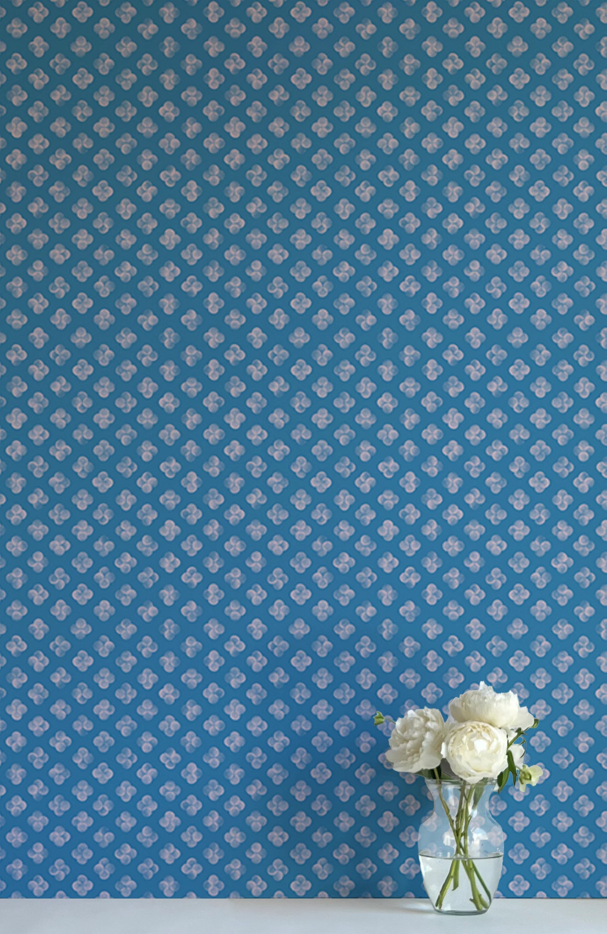 A vase of flowers stands in front of a wall covered in a minimalist quatrefoil pattern in light pink on a blue field.