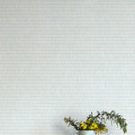 A vase of flowers stands in front of a wall covered in a textural grid print in shades of light blue and cream.