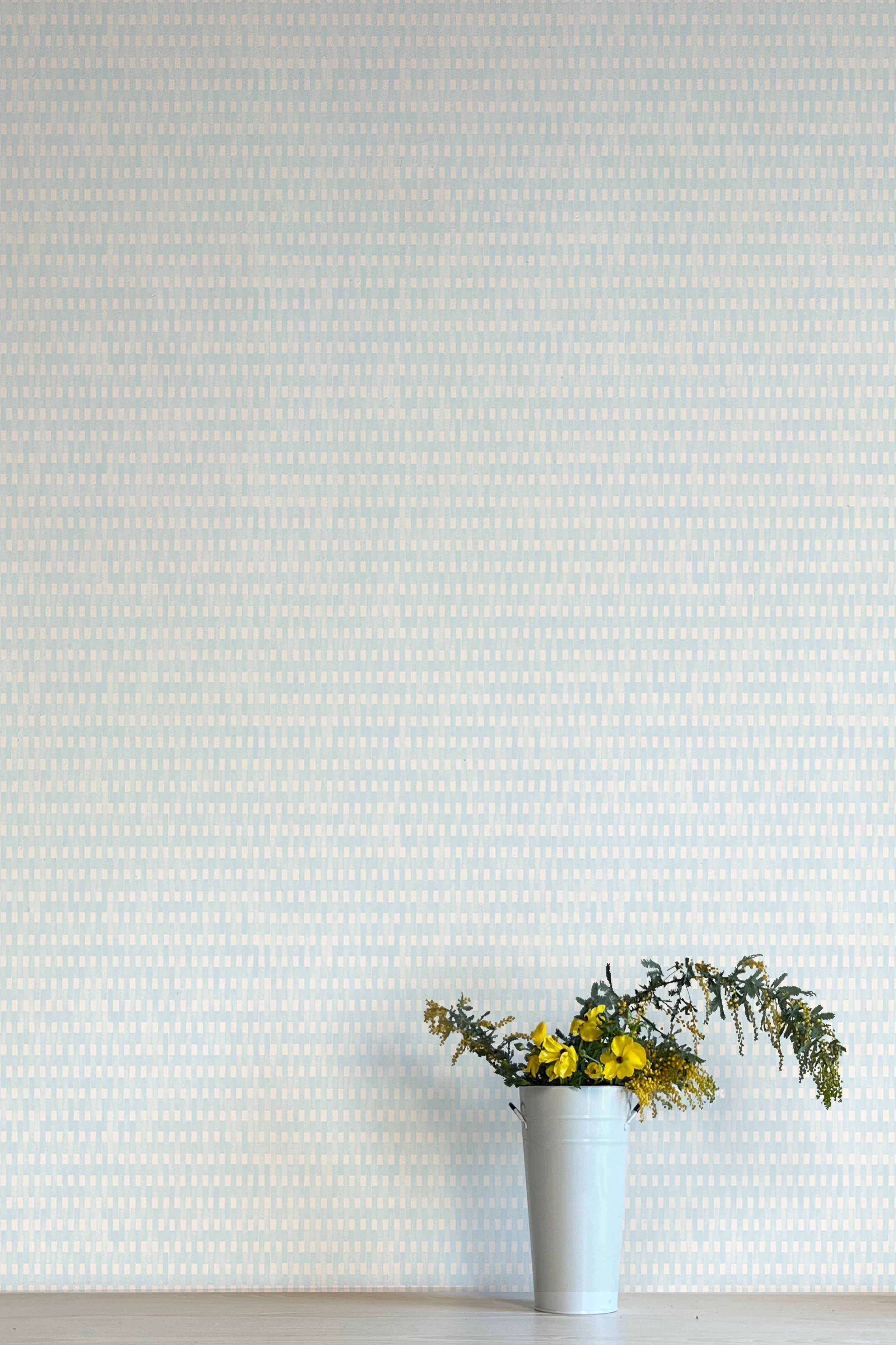 A vase of flowers stands in front of a wall covered in a textural grid print in shades of light blue and cream.