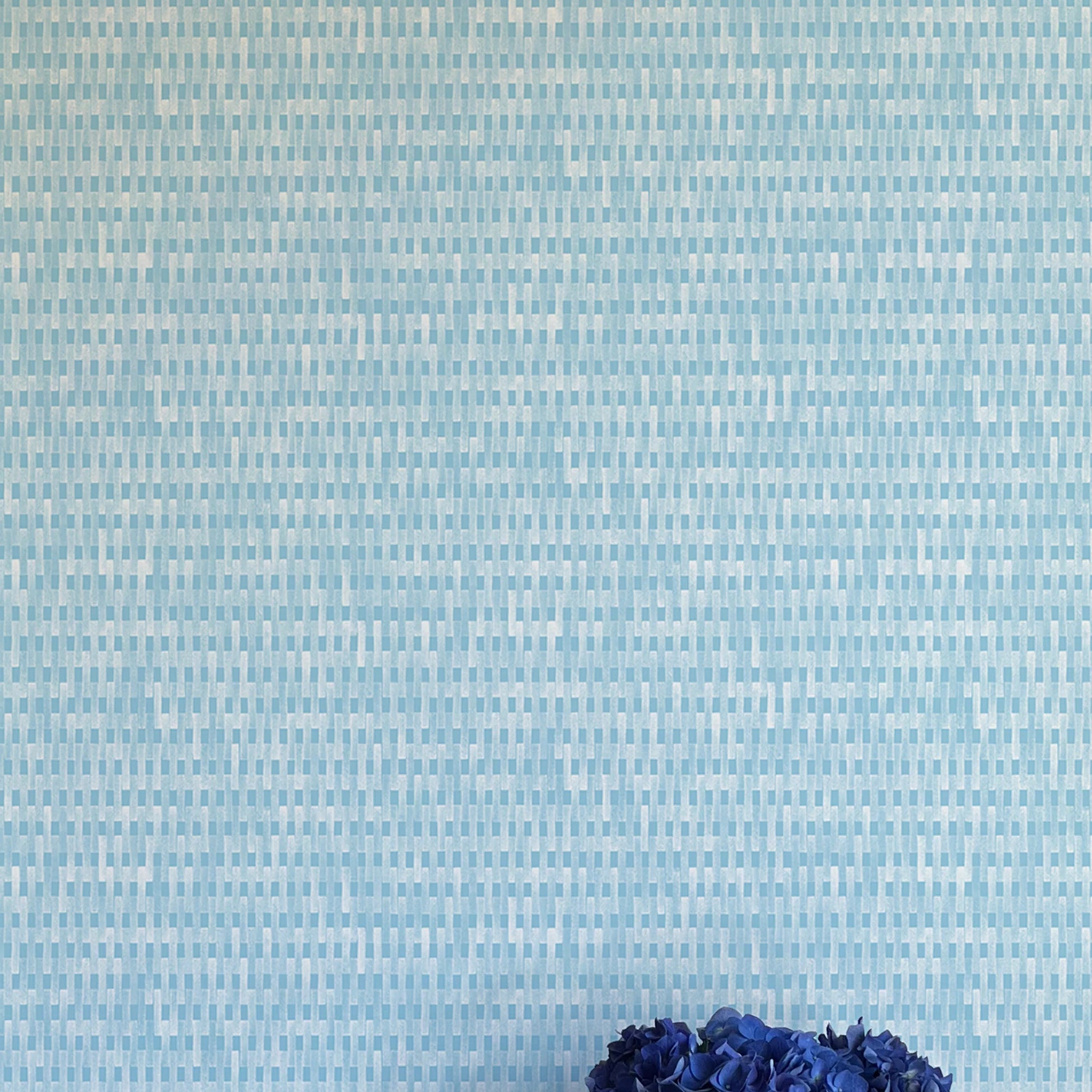 A vase of flowers stands in front of a wall covered in a textural grid print in shades of blue and cream.